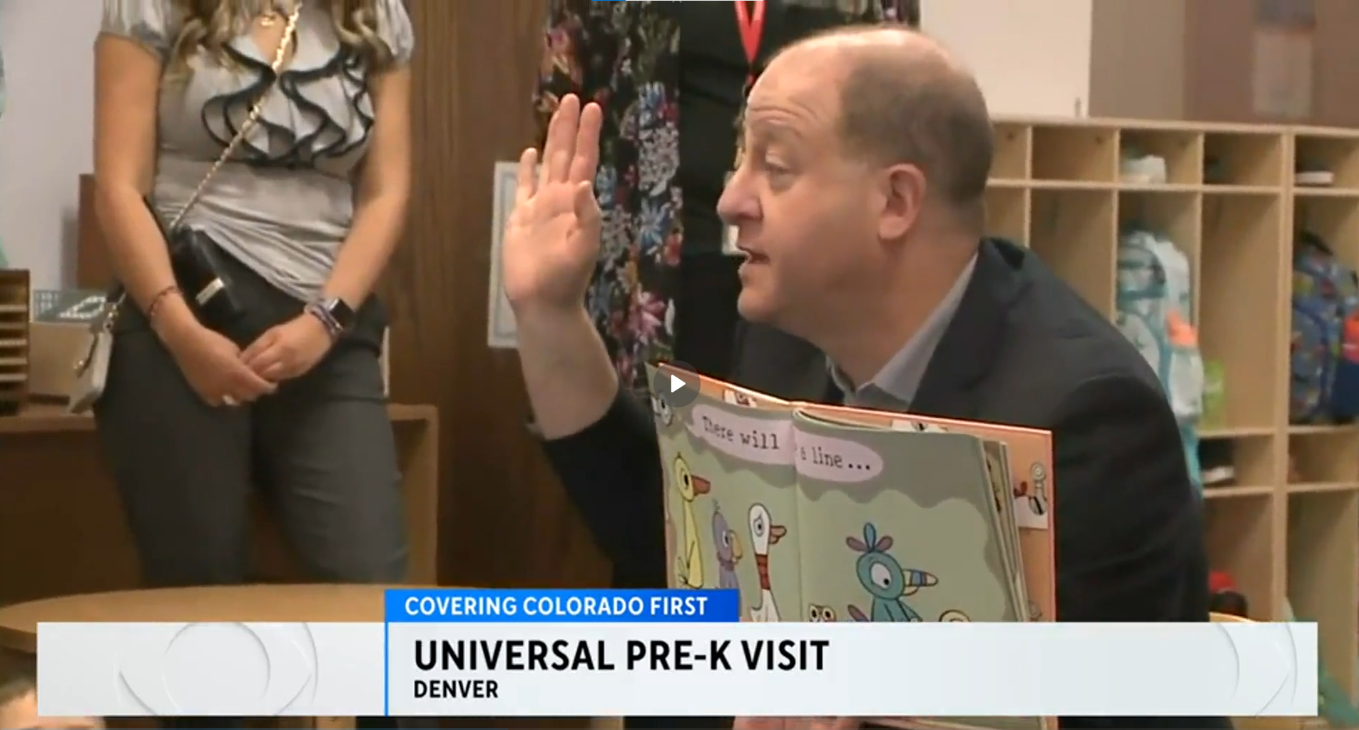 A person reads a childrens book to a group in a classroom. The book cover shows cartoon birds. Others stand in the background. The text overlay reads, UNIVERSAL PRE-K VISIT DENVER..