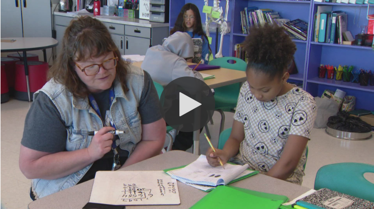 A teacher sits beside a student who is writing in a notebook. Other students work in the background. Classroom shelves with books and supplies are visible behind them.