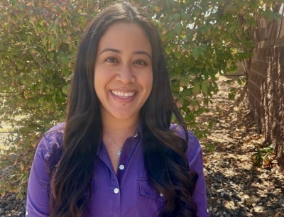 A woman with long dark hair, wearing a purple shirt, smiles while standing outside in front of leafy bushes and a wooden fence on a sunny day.