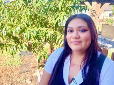 A person with long dark hair and purple streaks smiles outdoors, sitting near a leafy tree. They wear a white shirt, a black vest, and have visible face makeup. The background features a shaded wooden structure.