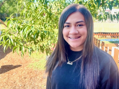 A young woman with straight brown hair, wearing a black top and a necklace, smiles outdoors near a leafy tree on a sunny day.