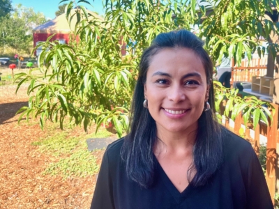 A woman with long dark hair, wearing a black shirt, smiles at the camera while standing outside in a sunny garden with green trees and a wooden fence in the background.