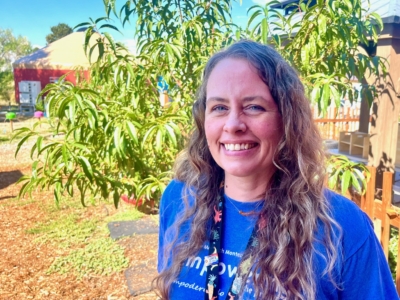 A woman with long wavy hair smiles while standing outside in front of a green leafy tree. She is wearing a blue T-shirt and a lanyard. The background includes a wooden fence and garden.