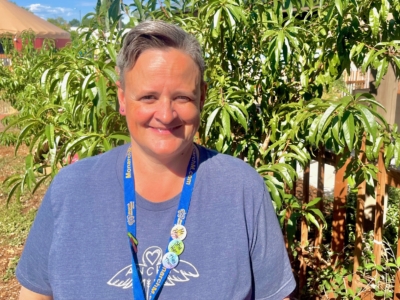 A person with short gray hair smiles outdoors, wearing a blue shirt and blue lanyard with pins, standing in front of green plants and a wooden fence under a sunny sky.
