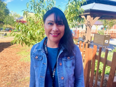A woman with long dark hair, wearing a denim jacket and lanyard, smiles outdoors in front of a wooden fence, trees, and a gazebo on a sunny day.