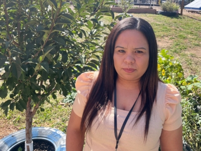 A woman with long dark hair, wearing a light peach top and a lanyard, stands outdoors near a leafy tree and greenery under bright sunlight.