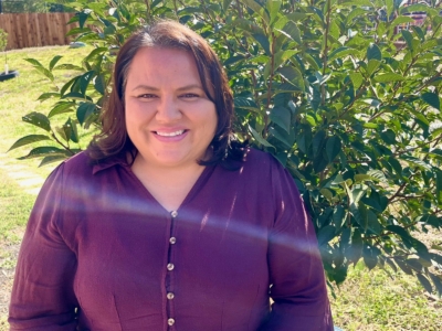 A woman wearing a maroon blouse smiles while standing outside in front of a large green bush, with sunlight creating a lens flare across the image.