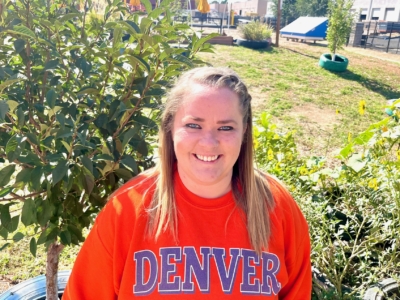 A person wearing an orange DENVER sweatshirt smiles outdoors in a garden with green plants and trees visible in the background.