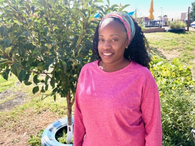 A woman wearing a pink shirt and colorful headband stands outdoors near a small tree, smiling at the camera on a sunny day.