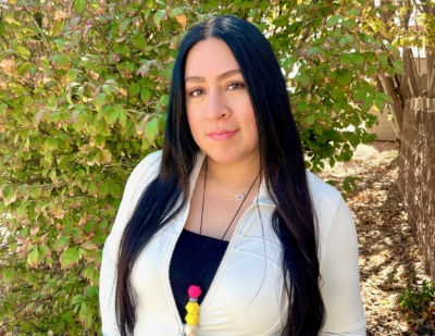 A woman with long dark hair stands outside in front of leafy green bushes, wearing a light jacket over a black top and a necklace with colorful pom-poms. Sunlight brightens the scene.