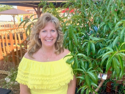 A woman with wavy blonde hair wearing a bright yellow off-the-shoulder top smiles while standing in front of green leafy plants at an outdoor market or garden area.