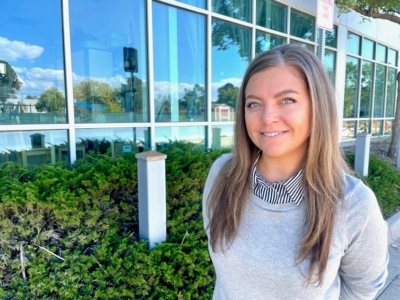 A woman with long brown hair, wearing a gray sweater and striped shirt, stands outside near green bushes and a glass building on a sunny day.