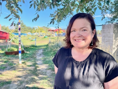 A woman with shoulder-length brown hair and a black shirt smiles outdoors near a garden with colorful poles, greenery, and a red structure in the background on a sunny day.