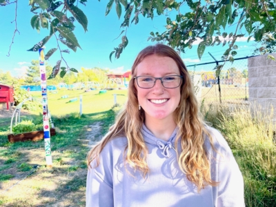 A young woman with glasses and long blonde hair smiles outdoors on a sunny day, standing in front of a colorful pole, fence, and grassy field with trees and a blue sky in the background.