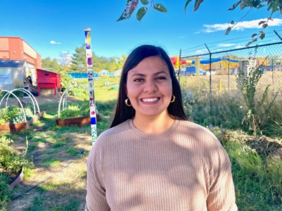 A woman with long dark hair, wearing a light brown sweater, smiles outdoors in a garden with colorful structures, plants, and a blue sky in the background.
