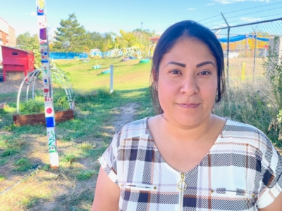 A woman with long dark hair stands in a sunny outdoor garden with colorful poles, playground equipment, and greenery visible in the background. She is wearing a plaid shirt and looking at the camera.