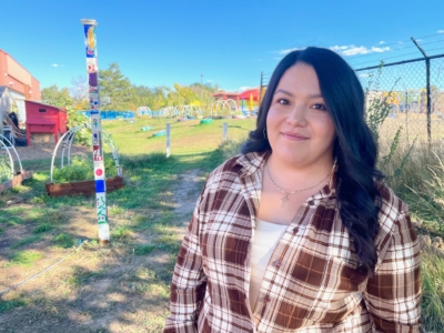 Woman with long dark hair and a plaid shirt stands outdoors in a sunny garden area, with a decorated pole and fence in the background.