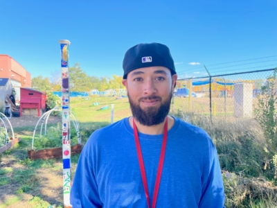 A man with a beard and black cap stands outside in a garden area, wearing a blue shirt and red lanyard. There is a colorful pole and wire fence in the background under a bright blue sky.