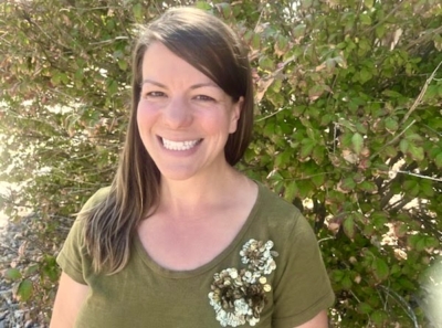 A woman with long brown hair, wearing an olive green shirt with floral decorations, smiles while standing outdoors in front of leafy green bushes under sunlight.