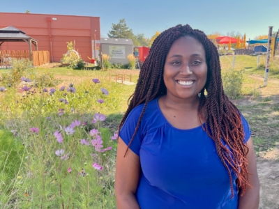 A woman in a blue shirt stands and smiles in a garden with purple flowers. There are buildings and outdoor structures in the background under a clear sky.