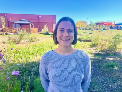 A woman with short brown hair, wearing a gray sweater, stands smiling in a grassy outdoor area with flowers, trees, and buildings in the background.