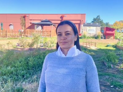 A woman with dark hair wearing a light gray sweater and white shirt stands in a garden with a red building and wooden fence in the background under a clear blue sky.
