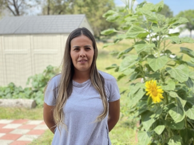 A woman with long brown hair, wearing a light gray shirt, stands in a garden beside a sunflower, with a shed and greenery in the background.