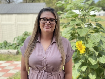 A woman with long straight hair and glasses stands outside in front of a sunflower and garden, wearing a light purple belted dress and smiling at the camera.