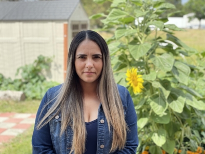 Woman with long, straight hair wearing a denim jacket stands in a garden with sunflowers and a shed in the background.