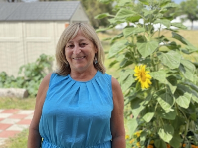 Woman in a sleeveless blue dress standing outside, smiling, with sunflowers and greenery in the background. A shed and tiled patio are visible behind her.