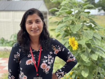 A woman with long dark hair, wearing a black floral top and a red lanyard, stands outdoors in front of a tall sunflower and a shed, smiling at the camera.