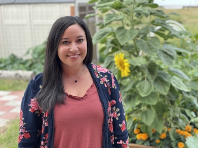 A woman with long dark hair, wearing a floral cardigan and rust-colored top, stands smiling in a garden with sunflowers and greenery in the background.