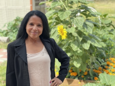 A woman with long dark hair stands outdoors in front of sunflowers and marigolds, wearing a black blazer over a light-colored top, smiling at the camera.
