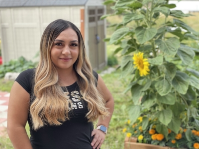 A woman with long, wavy hair stands in a garden next to a tall sunflower plant, smiling at the camera. A shed and greenery are visible in the background.