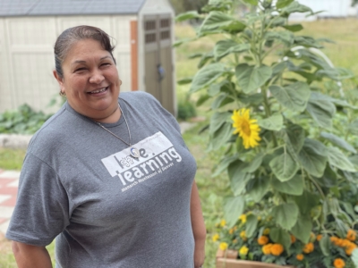 A woman wearing a gray learning t-shirt smiles while standing next to a tall sunflower in a garden with sheds and greenery in the background.