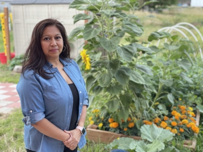 A woman stands in front of a lush garden with large sunflowers and marigolds, wearing a light blue button-up shirt over a black top, looking at the camera.