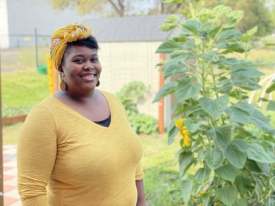 A smiling woman wearing a yellow headwrap and shirt stands next to tall sunflowers in a garden on a sunny day.