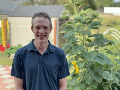 A person in a navy polo shirt smiles while standing in a garden with large green plants and sunflowers. There are sheds and a tiled patio in the background.