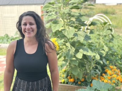 A woman with long brown hair, wearing a black sleeveless top and striped skirt, smiles while standing in a garden with sunflowers and yellow marigolds in the background.