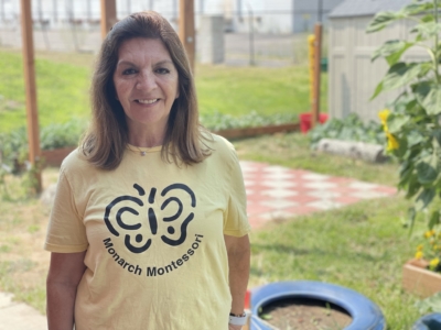 A woman with long brown hair smiles outdoors, wearing a yellow Monarch Montessori t-shirt. She stands in a garden area with green plants, a blue planter, and a checkered patio visible in the background.
