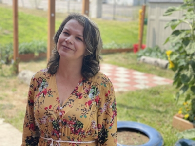 A woman in a yellow floral dress stands outside, smiling. There is greenery, a blue tire planter, and a checker-patterned area in the background. The scene appears to be in a garden or backyard.