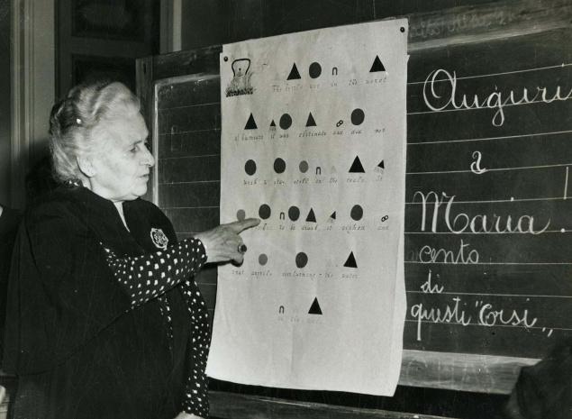 An older woman points to a chart with shapes on a blackboard. Italian writing and Auguri a Maria are on the board next to her. She appears to be teaching or explaining something.