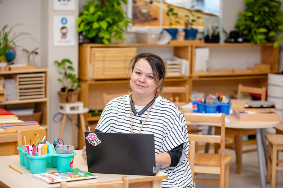 A woman sits at a desk in a bright classroom with plants, shelves, and art supplies. She smiles at the camera with a laptop and colorful stationery on the desk in front of her.