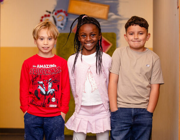 Three children stand side by side indoors, smiling at the camera. One wears a red Spider-Man shirt, another wears a pink outfit, and the third wears a beige shirt with blue jeans.