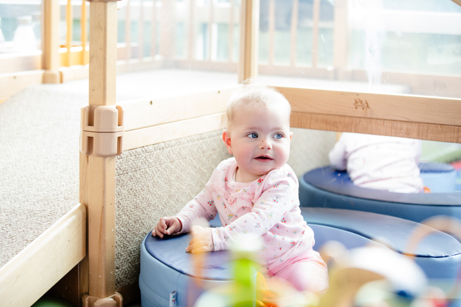 A baby in a pink outfit sits on a blue cushion in a bright play area, looking to the side. There are wooden structures and a mirror reflecting the babys back.