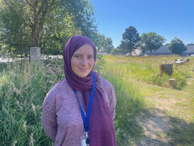 A woman wearing a purple hijab and a light purple top stands outside on a sunny day, smiling, with trees and grass in the background.