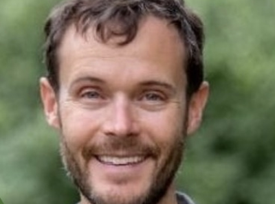 A man with short brown hair and a beard is smiling outdoors, with a background of blurred greenery.