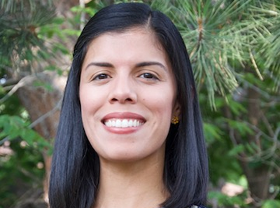 A woman with straight black hair, wearing a patterned top and small earrings, smiles while standing outdoors in front of green trees and foliage.
