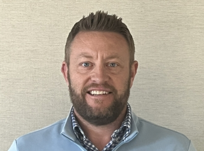 A man with short brown hair and a beard, wearing a light blue zip-up sweater over a blue checked shirt, smiles in front of a beige textured background.