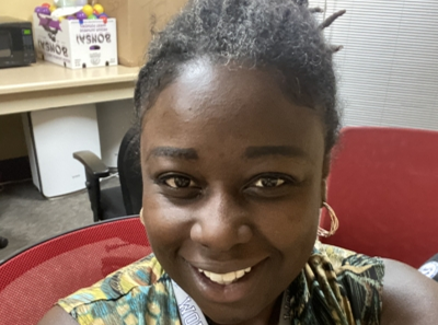 A woman with dark skin and short curly hair smiles at the camera while sitting in a red chair in an office setting with a fridge and boxes in the background.